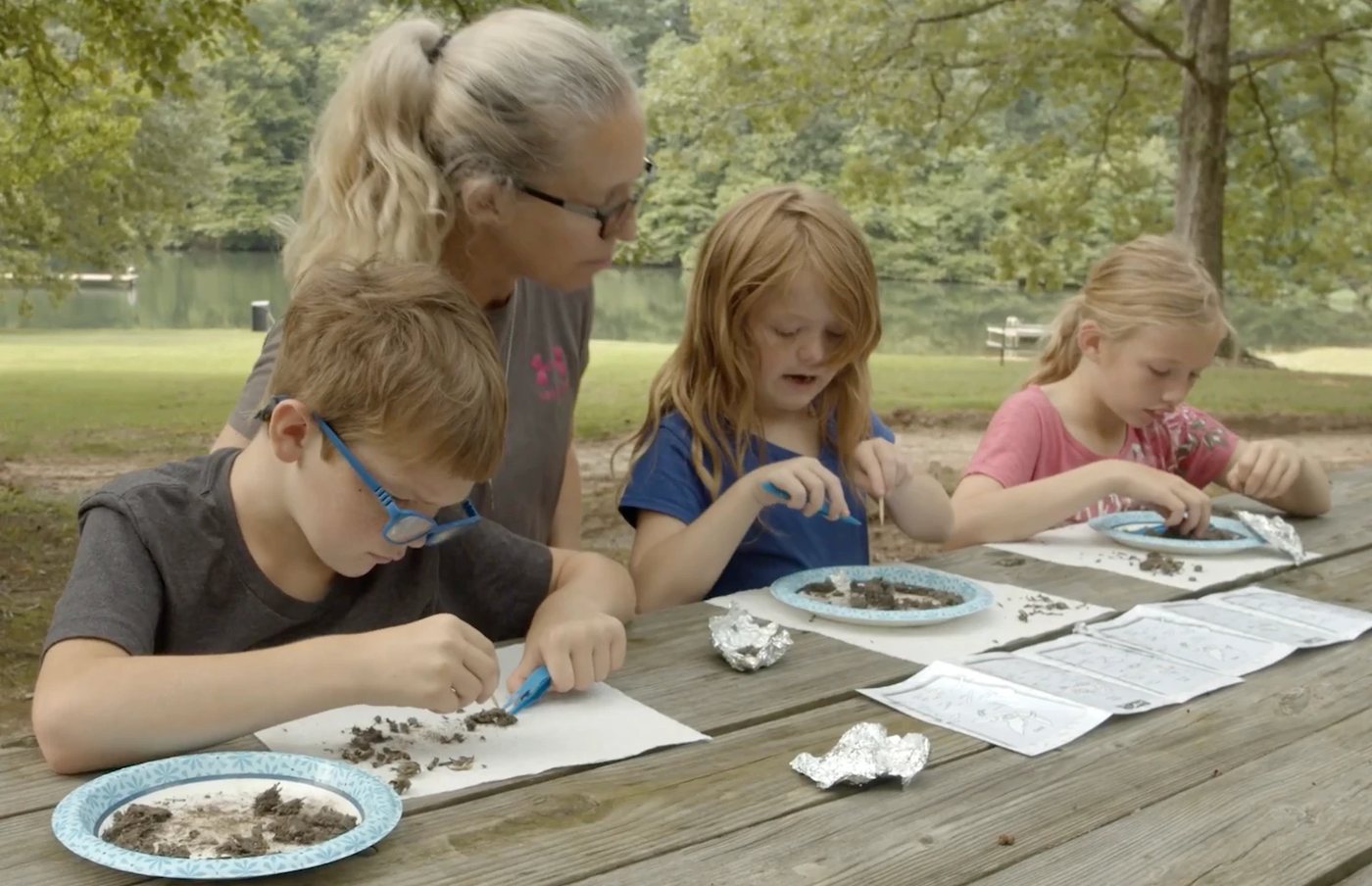 A teacher and three children dissecting owl pellets at a picnic table by a lake